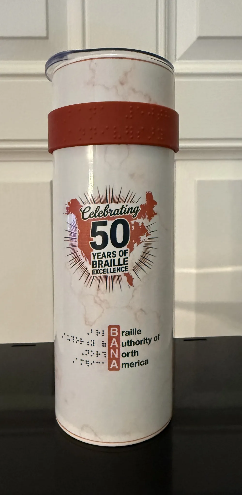 BANA 50th Anniversary braille tumbler, front: white marble finish with red resin grip band, reading &ldquo;Braille Rules. We Made Sure of It.&rdquo; above an illustration of a reader with a braille book, and the year 2026.